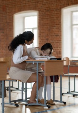 An adult sitting alongside a child at a classroom desk, offering quiet support